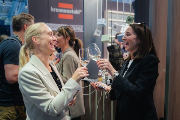 Zwei Frauen stoßen auf einem Messestand mit Weingläsern an; im Hintergrund hängen hugo! ONE Steckdosenwürfel.